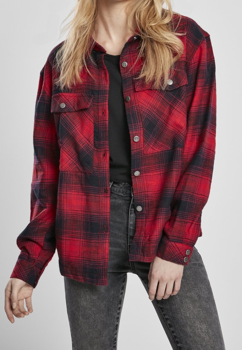 Kvinde iført en rød og sort ternet skjorte med knapper over en sort top og slidte sorte jeans, poserende med hænderne afslappet langs siden.