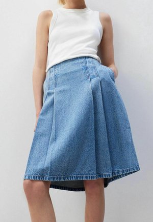 Femme portant un haut blanc sans manches et une jupe en jean bleue plissée, taille haute, longueur genou, les mains dans les poches.