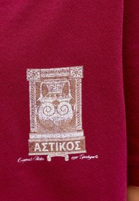 Sarkans kokvilnas T-krekls ar sīki izstrādātu grafiku bēšā krāsā, kas attēlo klasisko arhitektūru un satur Grieķu tekstu apakšā.