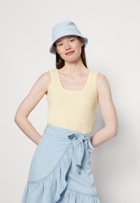 Top de malha amarelo clarinho com decote quadrado e bordas em forma de onda, combinado com uma saia envolvente de jeans azul claro e chapéu de balde.