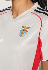Lichtgrijze voetbalshirt met een aansluitend ontwerp, rode accenten en een textuurpatroon. Inclusief een logo met een arendembleem op de borst.
