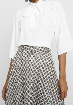 Kvinde iført en hvid bluse med en sløjfe foran ved kraven og et højtaljet, ankel-langt skørt med et brunt og hvidt ternet mønster.