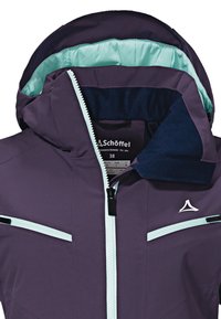 Fialová nepromokavá bunda s kapucí, s světle modrými zipy a detaily, a měkkou vnitřní podšívkou. Obsahuje logo značky a detaily označení.