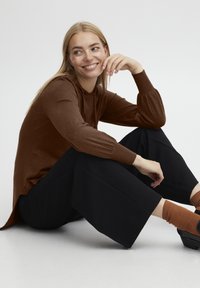 Sweat à capuche marron avec des manches côtelées, associé à un pantalon large noir. Le mannequin est assis, reposant sa main sur le genou. Une chaussette texturée est visible.