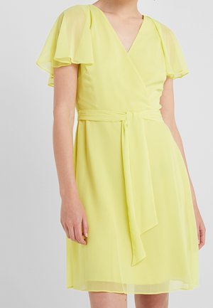 Donna che indossa un vestito avvolgente di colore giallo chiaro, lungo fino al ginocchio, con maniche a svolazzo e cintura in tessuto annodata in vita.