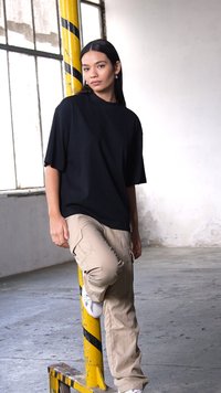 Černé oversized tričko s krátkým rukávem, béžové cargo kalhoty a bílé tenisky; opřený o sloup s žlutými pruhy v industriálním prostředí.