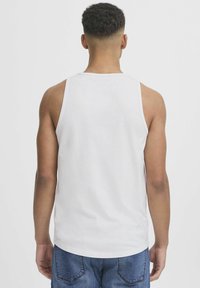 White cotton tank top with a round neckline and sleeveless design, paired with blue denim jeans. Back view shows smooth fabric texture.