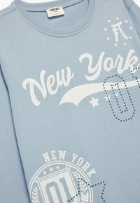 Jasnoniebieski sweter z białym napisem "Nowy Jork", graficznymi akcentami i okrągłym wzorem logo. Miękka tkanina z zaokrąglonym dekoltem.