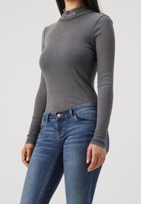 Grauer gerippter langärmeliger Oberteil mit einem Stehkragen und subtilen dekorativen Elementen, kombiniert mit taillierten blauen Jeans mit Knopfverschluss und Taschen.