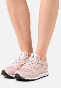 Zapatillas de deporte de color rosa claro, fabricadas en ante y malla, con un talón blanco. Presentan un logo en la lengüeta, cordones redondos y una suela de goma negra.