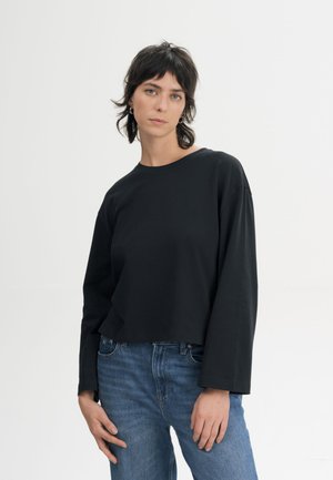 Vrouw met kort donker haar, gekleed in een zwarte top met lange mouwen en een blauwe jeans, staand tegen een effen lichte achtergrond.