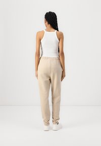 Beige trainingsbroek met elastische tailleband en boorden, gedragen met een witte tanktop. De stof lijkt zacht en glad, zonder zichtbare patronen.