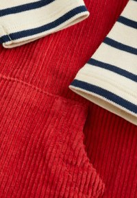 Roter Cordstoff mit vertikalen Rillen, kombiniert mit cremefarbener Stoff, der navyfarbene horizontale Streifen an den Bündchen aufweist. Sichtbare detaillierte Nähte.