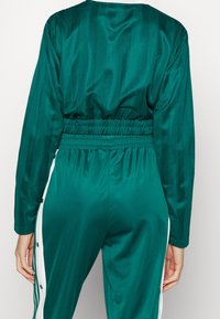 Teal satin träningsoverall med långa ärmar, samlad midja och vita sidoknappar. Har vertikala ränder för ökad textur.