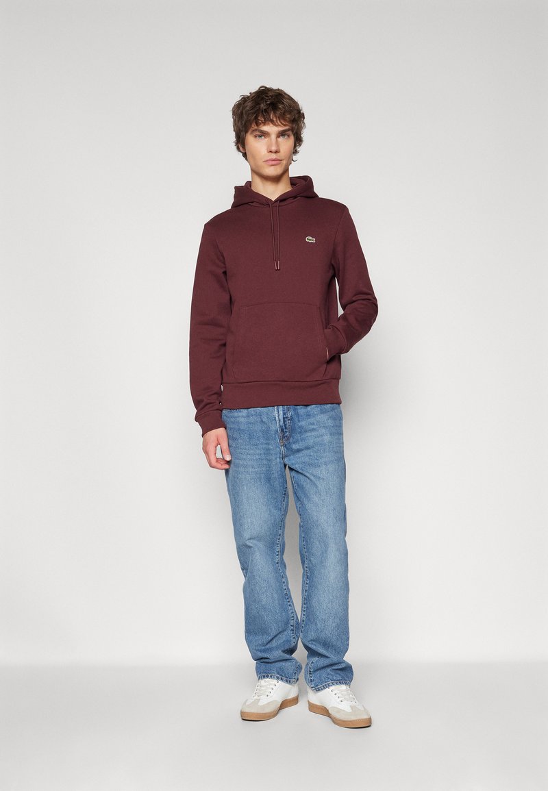 Burgunderfarbener Kapuzenpullover mit einer Kängurutasche, Kapuze mit Zugband und einem grünen Logo. Getragen mit hellblauen Jeans und weißen Sneakern.