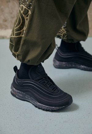 Sneakers sportive nere con suole ad aria visibili indossate con calzini neri e pantaloni verde oliva con motivi beige, in piedi su un pavimento chiaro macchiato.