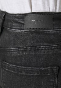 Los vaqueros de mezclilla negros presentan una textura suave con un brillo sutil, un parche de cuero negro con la marca en la cintura y costuras clásicas en los bolsillos.