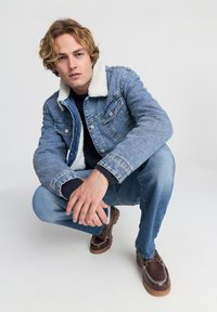 Giovane uomo con capelli biondi ondulati in posizione accovacciata, indossando una giacca di denim foderata in shearling blu, jeans blu e scarpe di pelle marroni.