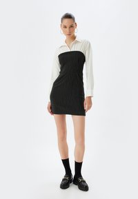 Schwarzes, gestreiftes Kleid mit einem weißen, kragenlosen Langarmshirt, taillierter Schnitt, knielang, kombiniert mit schwarzen Plateauschuhen und Socken.