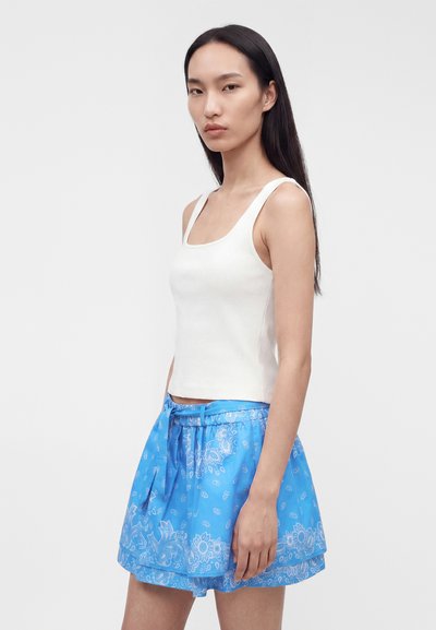 Γυναίκα με μακριά σκούρα μαλλιά, που φοράει λευκό τοπ χωρίς μανίκια και μπλε φούστα με σχέδια και ψηλή μέση με ζώνη, σε απλό φόντο.