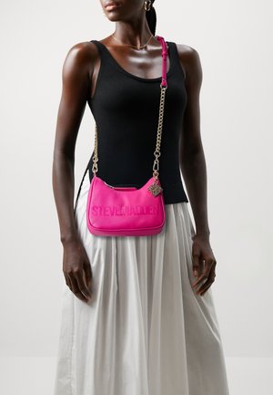 Woman wearing black tank top and white skirt, carrying bright pink Steve Madden crossbody bag with gold chain strap.