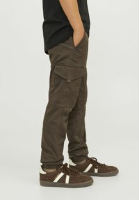 Tmavohnedé cargo nohavice s viacerými vreckami a voľným strihom, skombinované s hnedými teniskami so bielymi pruhmi a textúrovaným vrchom.