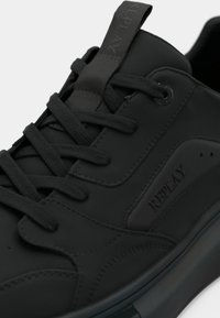Baskets noires en matériau synthétique lisse, avec un bout arrondi, des lacets plats, un logo discret et une semelle texturée au design minimal.