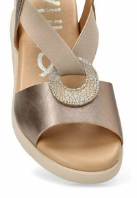 Sandal de punta abierta con parte superior de bronce metálico, tira elástica en cruz, acento decorativo circular y suela en tono marrón claro.