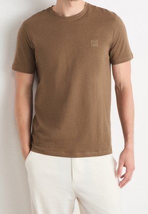 Homme portant un t-shirt marron uni à manches courtes et col rond avec un petit logo carré sur la poitrine, associé à un pantalon blanc.
