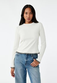 Langarm-T-Shirt aus weißem Rippmaterial mit gekürztem Saum und schwarzem Spitzenrand; kombiniert mit hochgeschnittenen hellblauen Denim-Jeans.