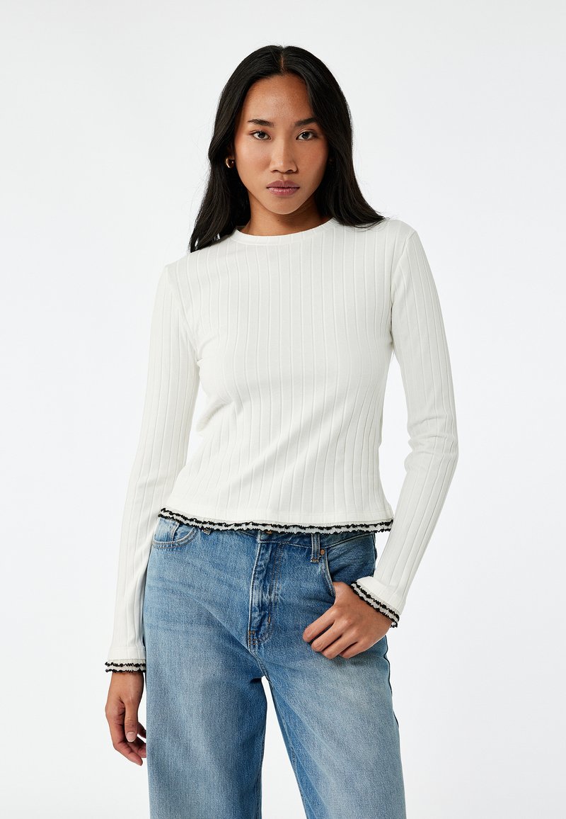 Langarm-T-Shirt aus weißem Rippmaterial mit gekürztem Saum und schwarzem Spitzenrand; kombiniert mit hochgeschnittenen hellblauen Denim-Jeans.