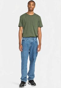 Groen T-shirt met korte mouwen en een borstzak, gecombineerd met lichtblauwe spijkerbroek en zwarte sneakers, staand tegen een neutrale achtergrond.