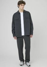 Chemise grise à manches longues avec deux poches poitrine, portée sur un T-shirt blanc et un pantalon gris assorti. Le modèle porte des baskets grises et argentées.
