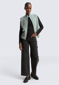 Veste tricotée vert clair avec col haut portée sur un haut noir à manches longues, associée à un jean large foncé et des chaussures plateformes noires.