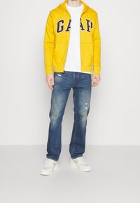 Sudadera amarilla con cremallera y logo "GAP", camiseta blanca, vaqueros desgastados azules y zapatillas blancas; ajuste casual con tejido texturizado.