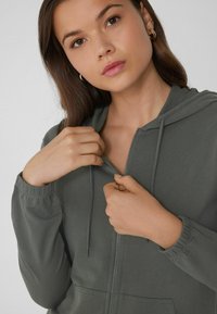 Mujer joven con pelo castaño que lleva pendientes de aro dorados, ajustando la cremallera de una sudadera con capucha gris oscuro y puños elásticos.