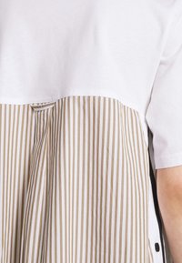 Valkoinen puuvillapaita, jossa on vaalean beige ja valkoinen raidallinen alaosa, suora leikkaus ja sivupainonapit.