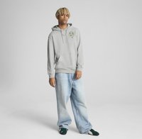 Grå hættetrøje med frontlomme og et grønt logo-patch. Løs pasform med lyseblå jeans og mørkegrønne sko fuldender outfittet.