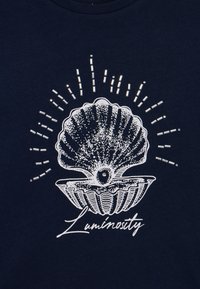 T-shirt azul-marinho com um gráfico de concha branca acompanhado por raios e realces, acompanhado pelo texto "Luminosidade" em uma fonte cursiva.