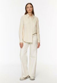 Chemise crème à manches longues, coupe décontractée ; associée à un pantalon large ivoire et des chaussures à plateforme beiges.