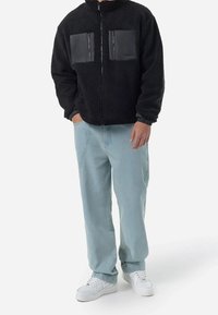 Mężczyzna w czarnej polarowej kurtce z dwiema przednimi kieszeniami na zamek, w jasnoniebieskich dżinsach i białych sneakersach, stojący z jedną ręką w kieszeni.