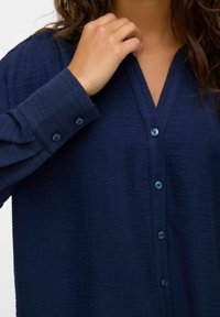 Camicia azzurra navy con bottoni, caratterizzata da un tessuto testurizzato, scollatura a V, maniche lunghe con dettagli a bottoni e bottoni coordinati lungo il davanti.