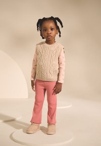 Bēšs adīts veste pār pasteļrozā ziedu raksta garroku topu, kombinēts ar rozā riboto biksēm ar volānu apakšmalu un bēšiem ziedu raksta zābakiem.