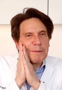 Un homme portant une blouse de laboratoire blanche sur une chemise rayée bleue et blanche, les mains sur le visage, avec des cheveux foncés et un fond neutre.