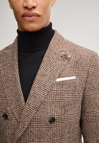 Dubbel gevoerde bruine geblokte blazer met zwarte knopen, een witte pochet en een gestructureerde zwarte coltrui eronder.