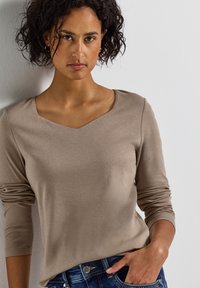 Haut beige à manches longues avec un décolleté en V et des manches ajustées, fabriqué en tissu doux, associé à un jean bleu avec un bouton argenté en accent.