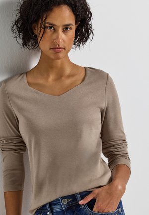 Beige langærmet top med V-hals og tætsiddende ærmer, lavet af blødt stof, parret med blå jeans med en sølvfarvet knapdetalje.