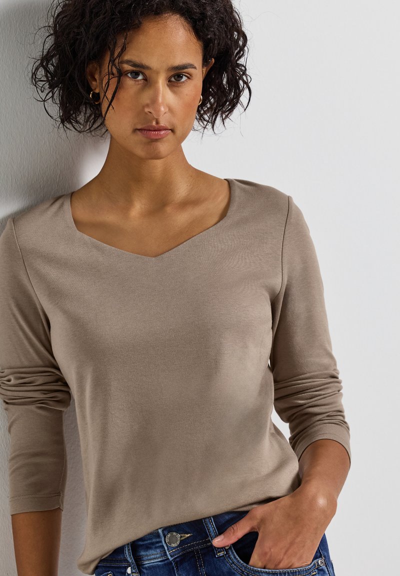 Haut beige à manches longues avec un décolleté en V et des manches ajustées, fabriqué en tissu doux, associé à un jean bleu avec un bouton argenté en accent.