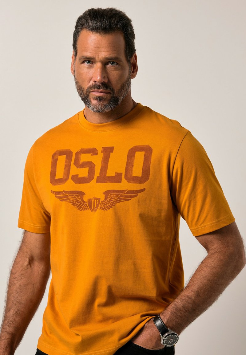 Oranžové bavlněné tričko s tučným hnědým nápisem "OSLO" a grafikou křídel. Krátké rukávy a kulatý výstřih. Obsahuje jemnou texturu.