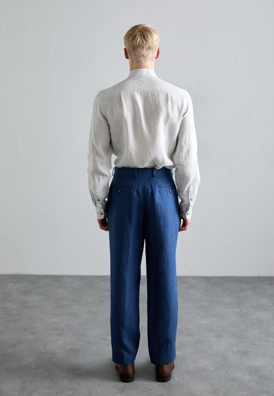 Camisa de lino gris claro con puños abotonados, combinada con pantalones de tiro alto azul oscuro. Textura suave, ajuste entallado, zapatos marrones visibles.